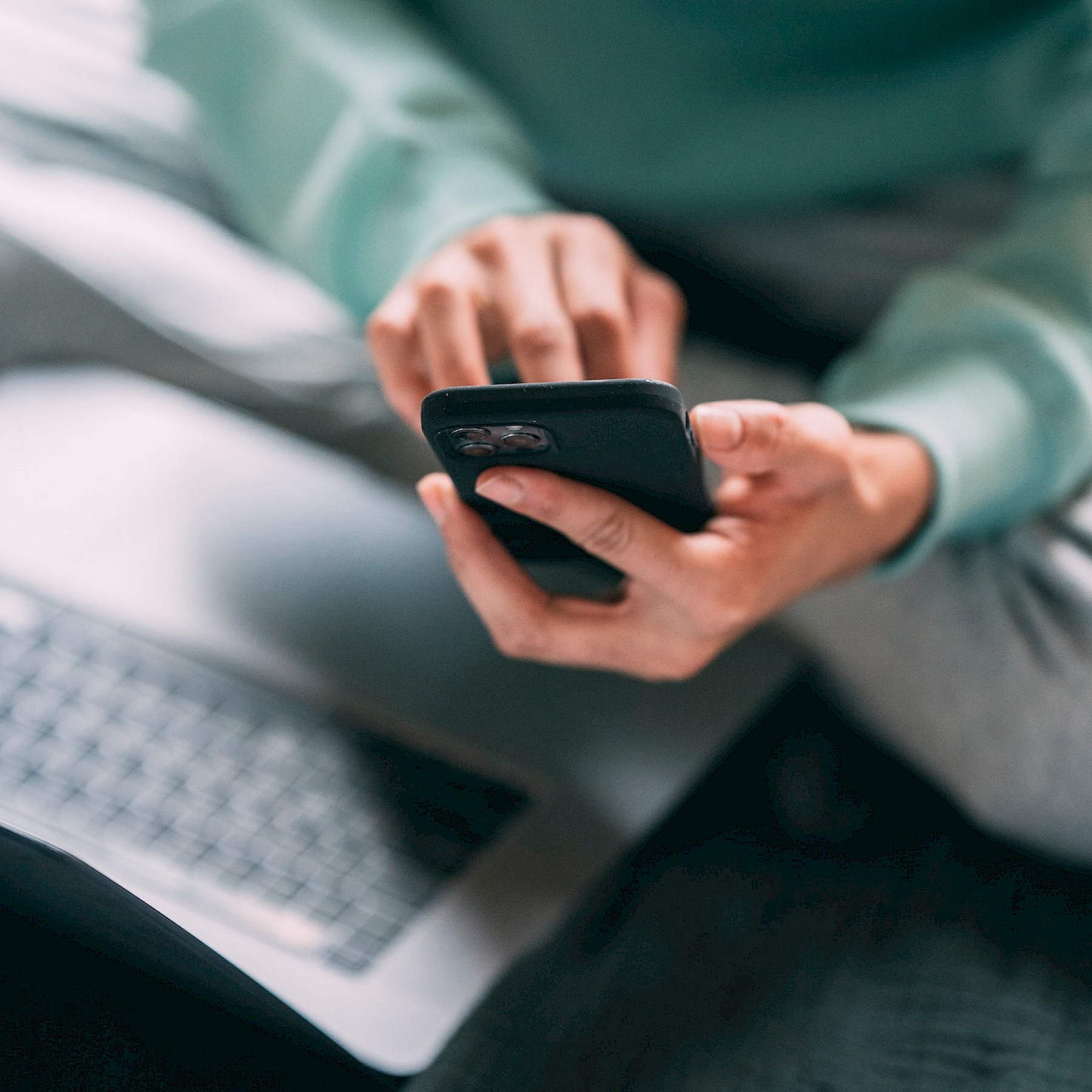 Ihminen istuu sängyllä edessään tietokone ja kädessään puhelin. Kasvoja ei näy. Näkökulma yläviistosta.